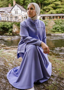 Woman in a purple dress sitting on a log by a stream with trees and a building in the background