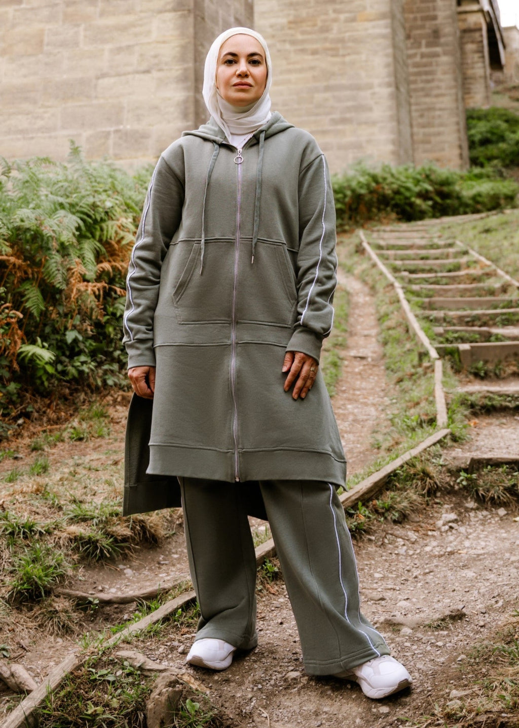 Person wearing a gray outfit standing on steps near a stone bridge with greenery.
