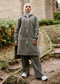 Person wearing a gray outfit standing on steps near a stone bridge with greenery.